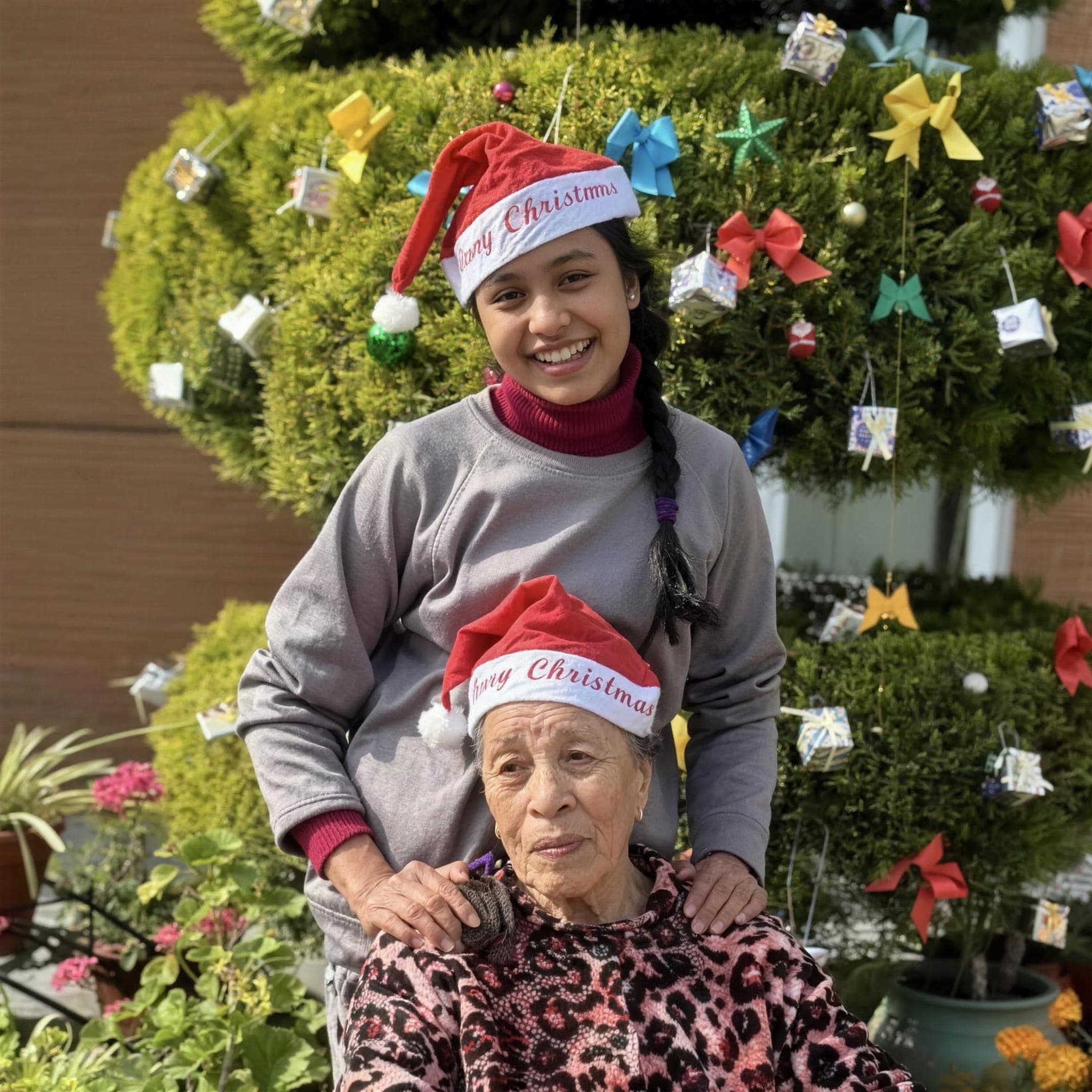 A Naman nurse and an elderly resident in matching Santa hats beside the Christmas tree