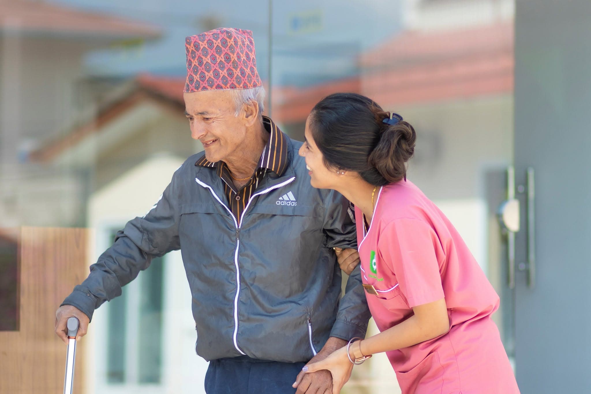 A Naman nurse walking alongside an elderly resident in a Nepali dhaka topi — a nurse companion for hospital visits