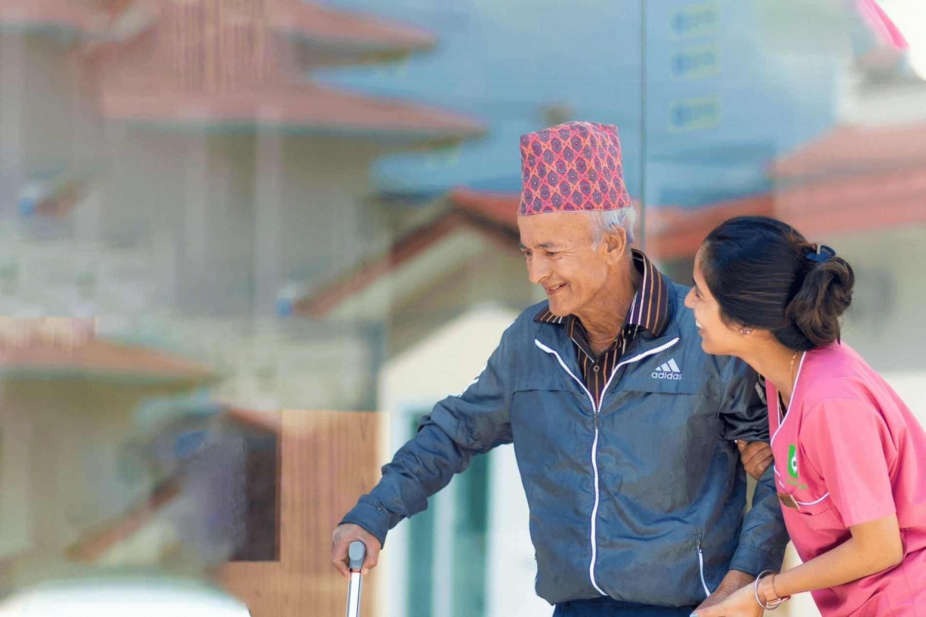 A Naman caregiver walking with an elderly resident wearing a dhaka topi, with our ward in the background