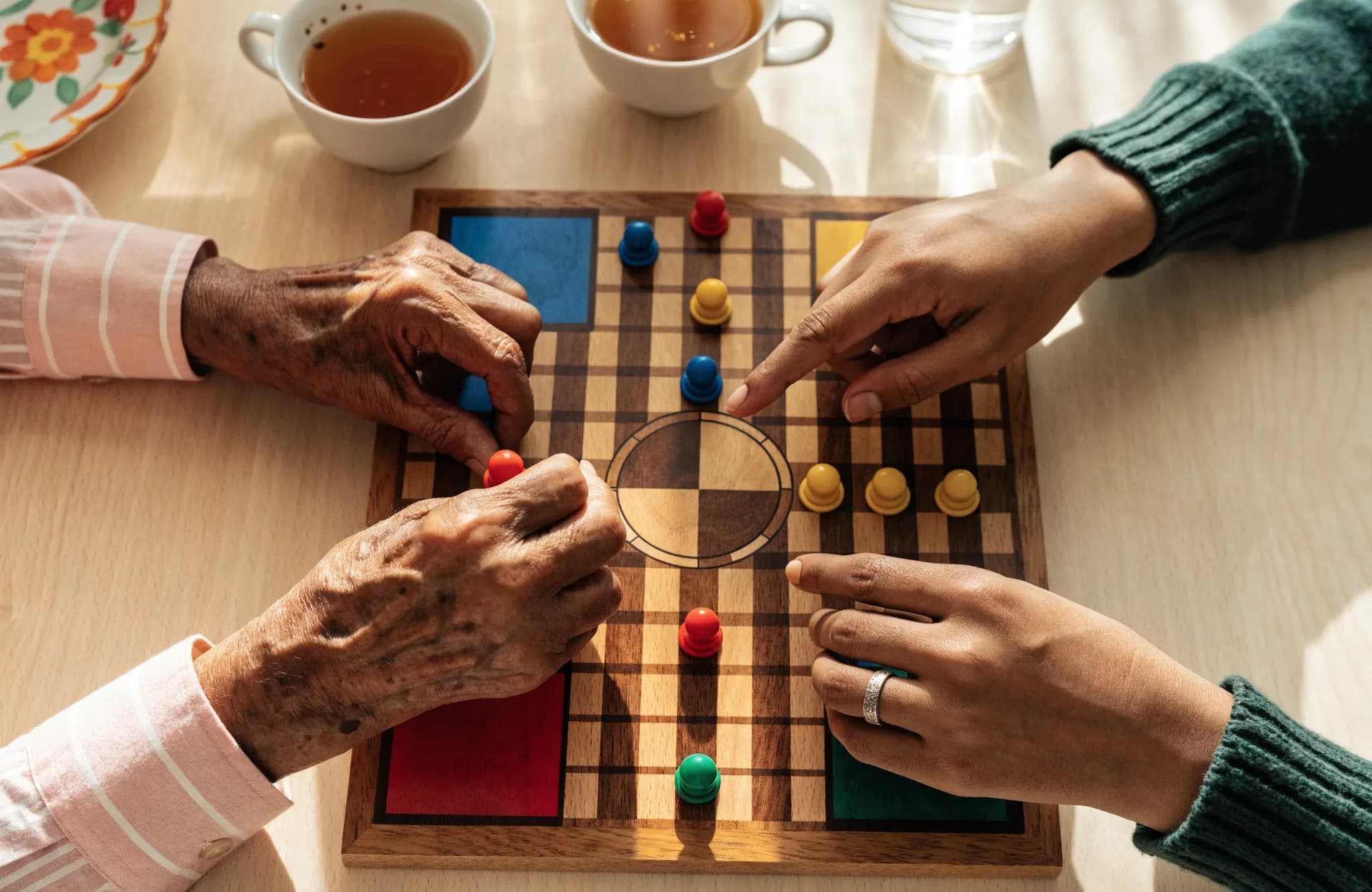 Overhead view of older and younger hands playing a wooden ludo board game over cups of tea
