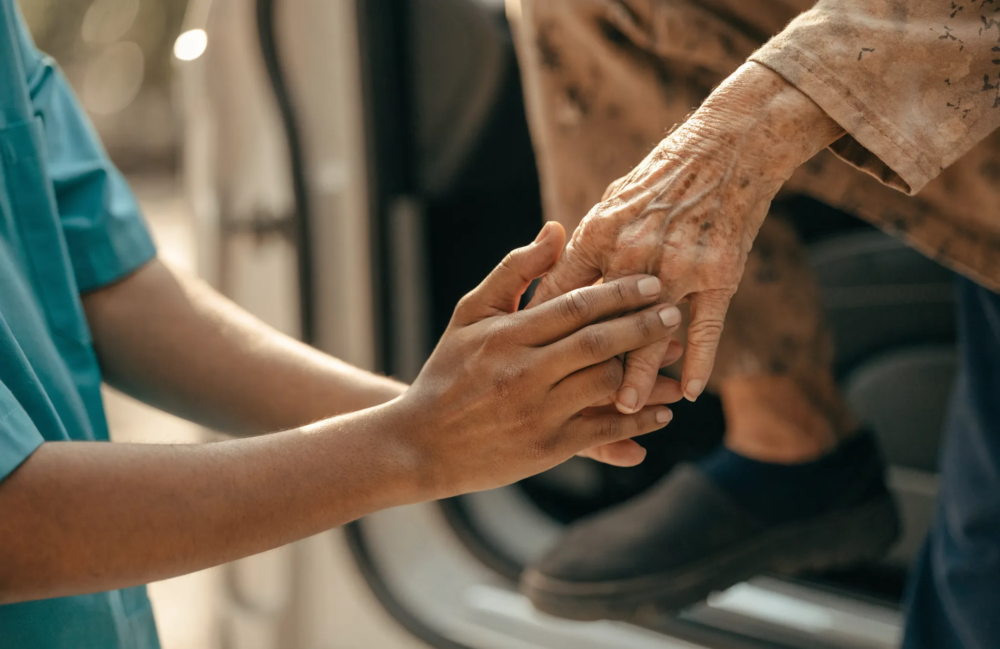 A caregiver's hand gently steadying an elderly person's hand beside a car door